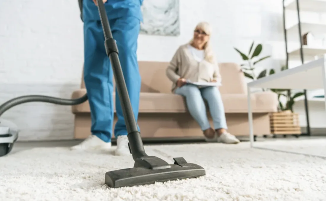 Auxiliar de Magik Clean aspirando la alfombra de un living mientras la clienta descansa en el sofá